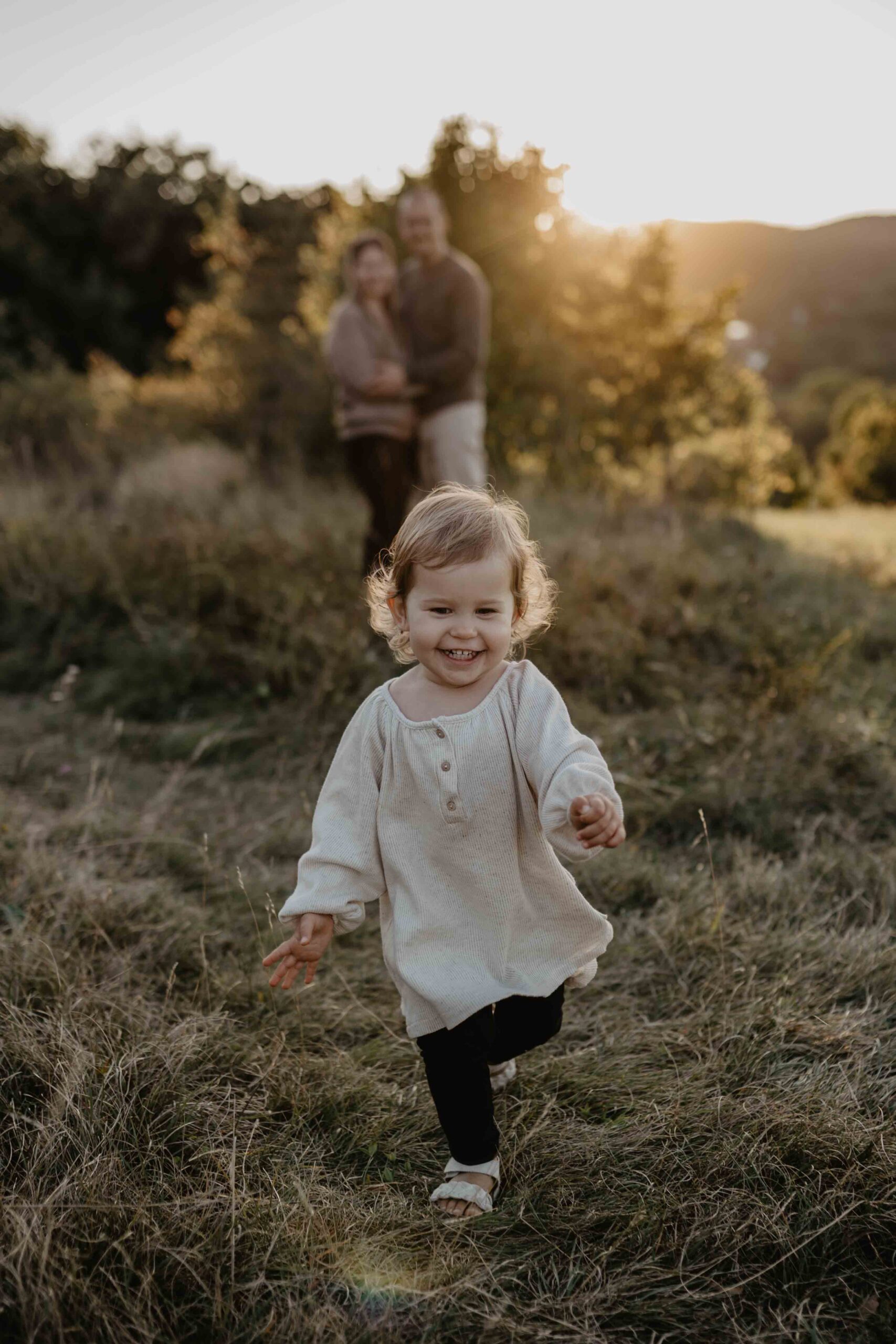 Während dem Fotoshooting in Mödling, Wien Umgebung, läuft die Tochter auf die Kamera zu, während die Eltern im unscharfen Hintergrund grinsen.