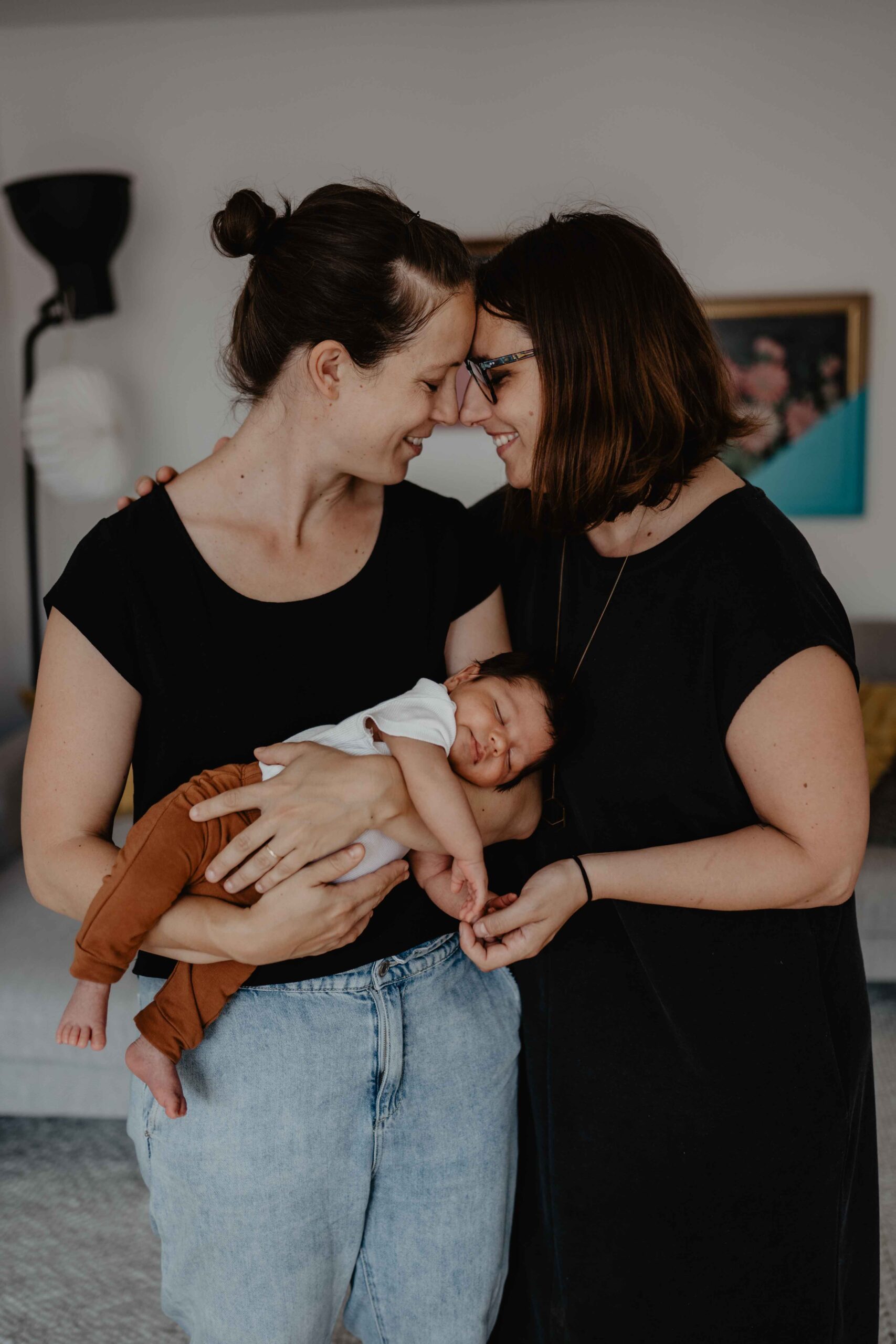 2 Mamas stehen als Regenbogenfamilie gemeinsam mit ihrem Sohn da, Stirn an Stirn und grinsen. Der Sohn schläft.