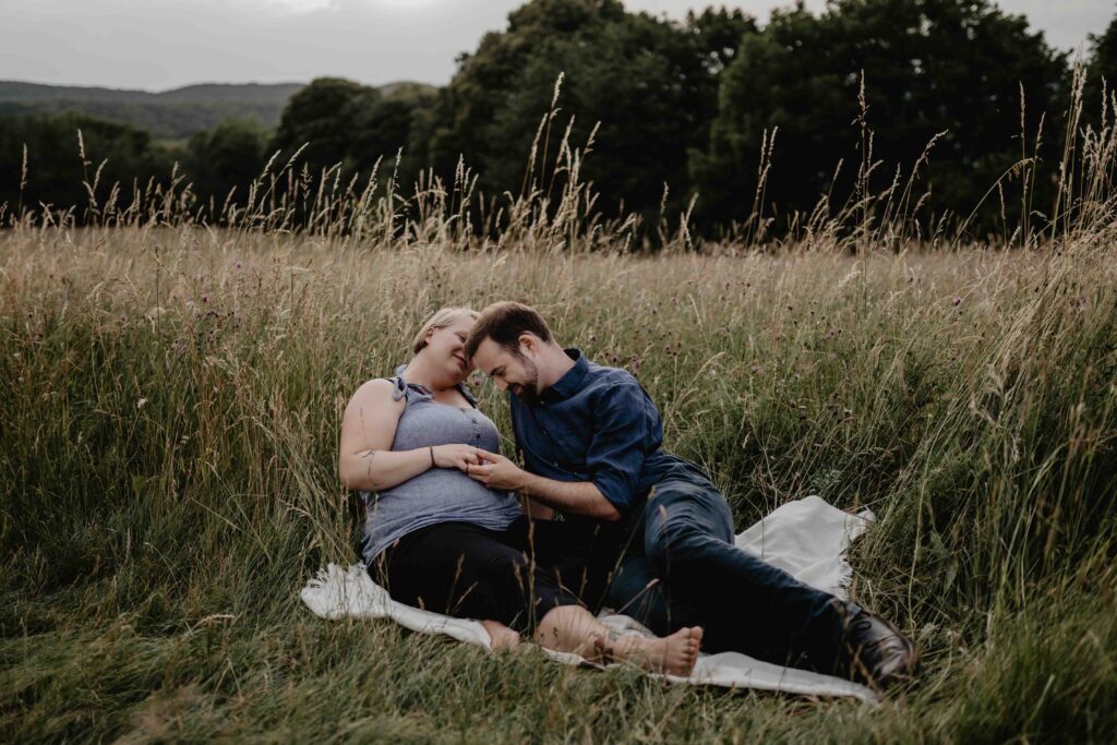 Ein schwangeres Paar sitzt in der Wiese und streichelt den Babybauch. Eine Babybauchfotografin hält dies für die drei fest.