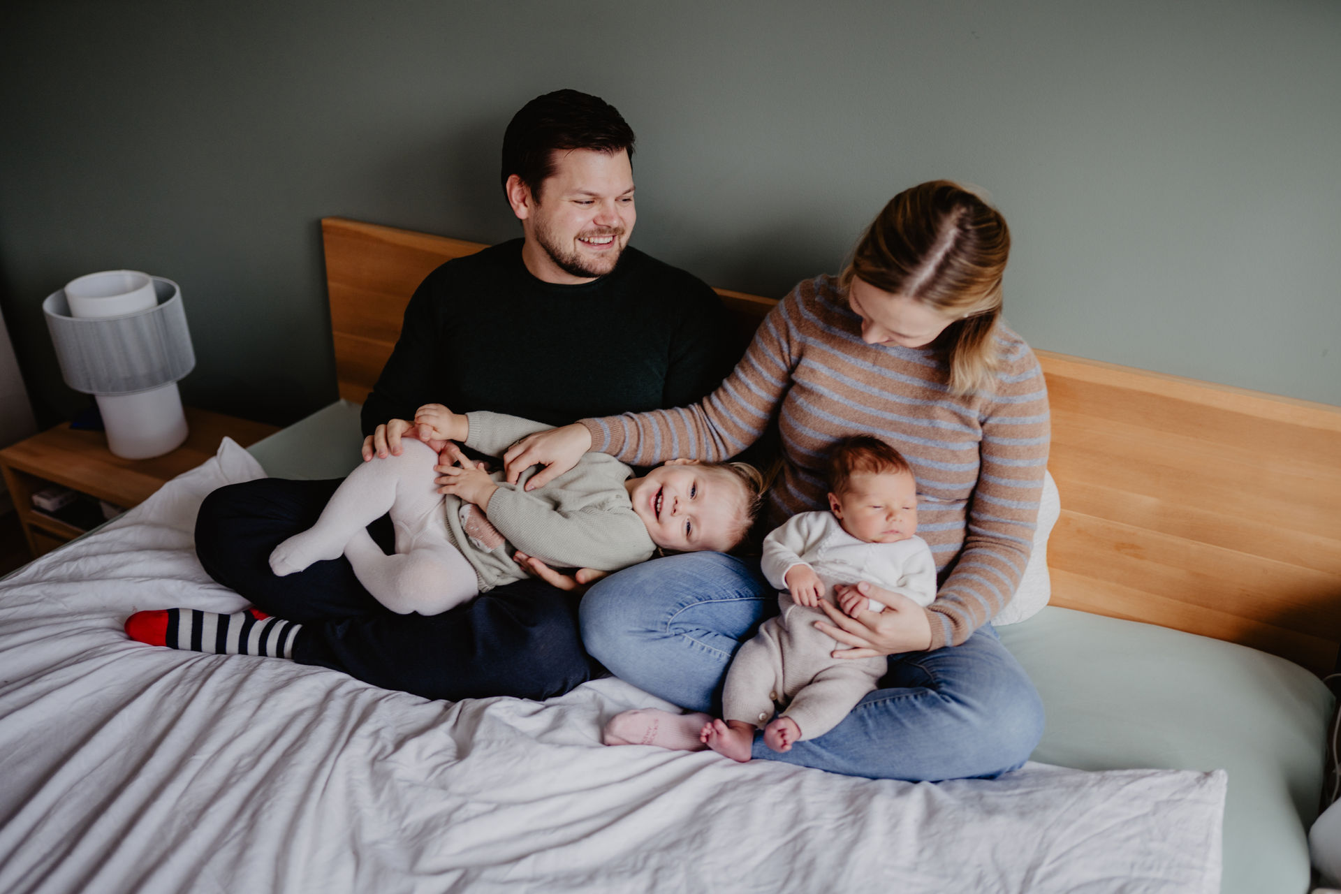 Die Familie sitzt auf ihrem Bett und es entstehen natürliche Newbornfotos in Wien und Umgebung. Alle haben ein großes Grinsen im Gesicht.
